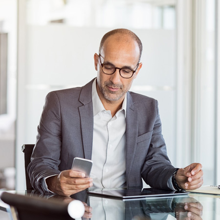 Mature business man in formal clothing wearing spectacles using mobile phone. Serious businessman using smartphone and digital tablet at work. Manager in suit using cellphone in a modern office.